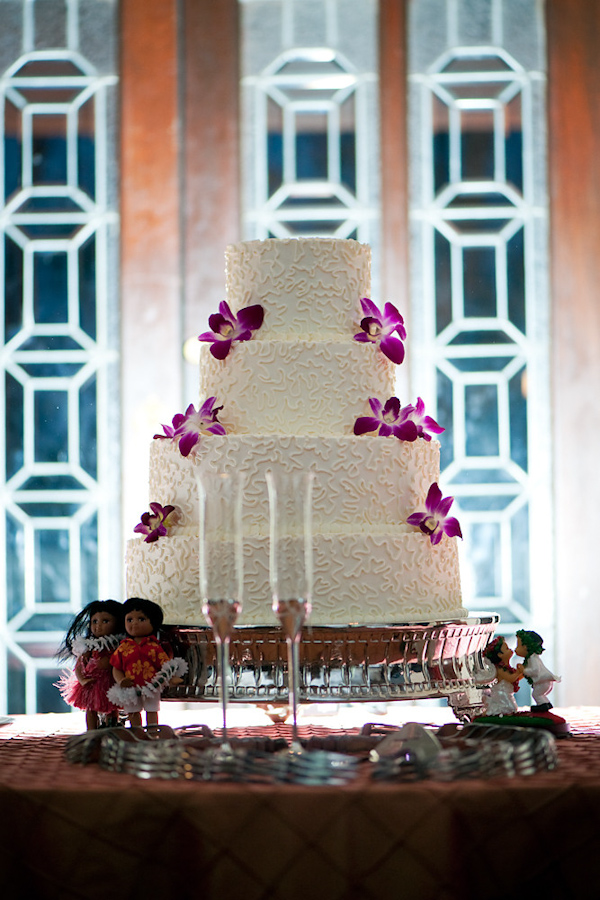 White four tiered wedding cake with purple tropical floral accents and a pair of hawaiian dolls - photo by Houston based wedding photographer Adam Nyholt
